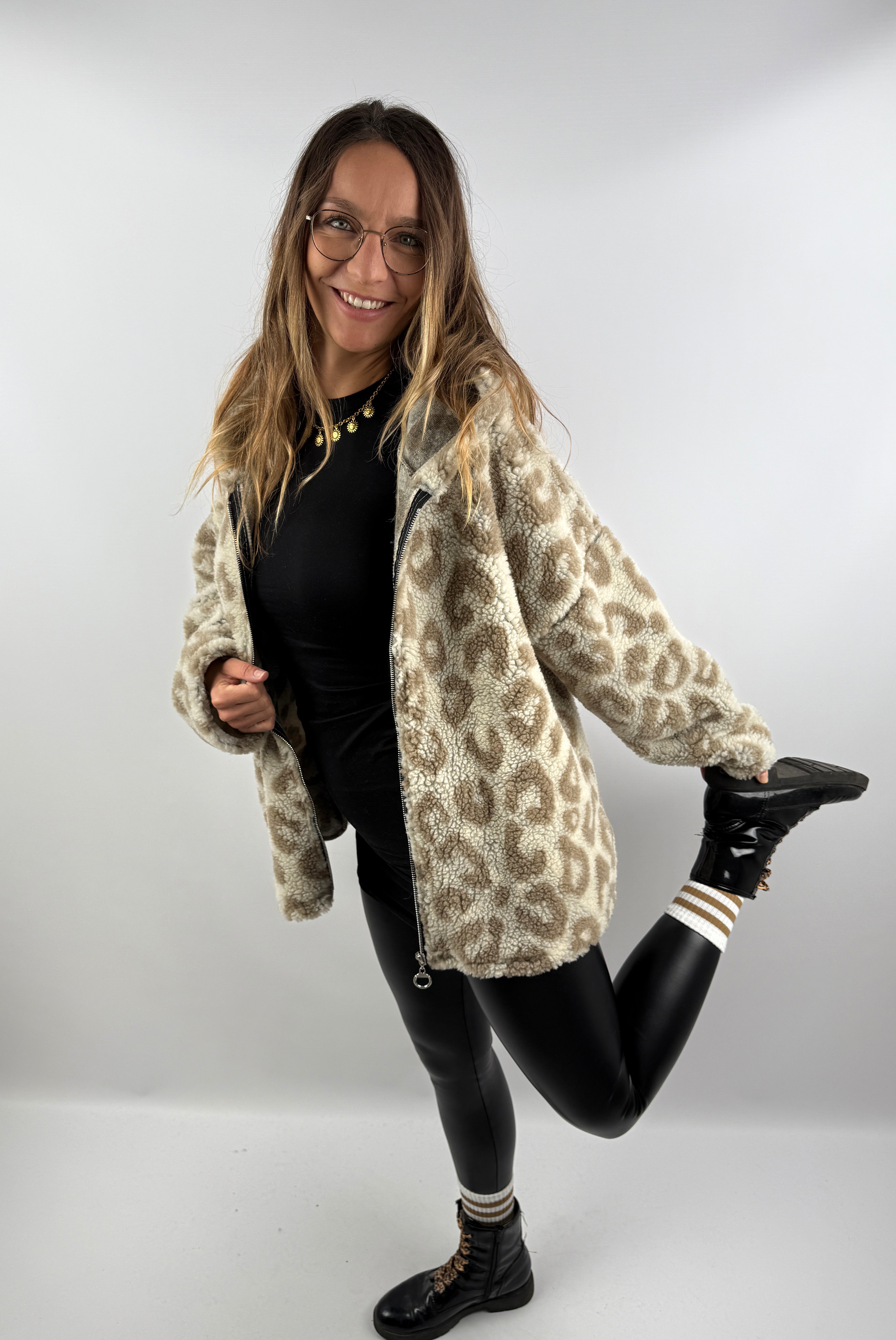 Frau posiert fröhlich in einer beigen Leo Teddy Jacke mit Leo-Muster und Kapuze, kombiniert mit schwarzem Outfit.