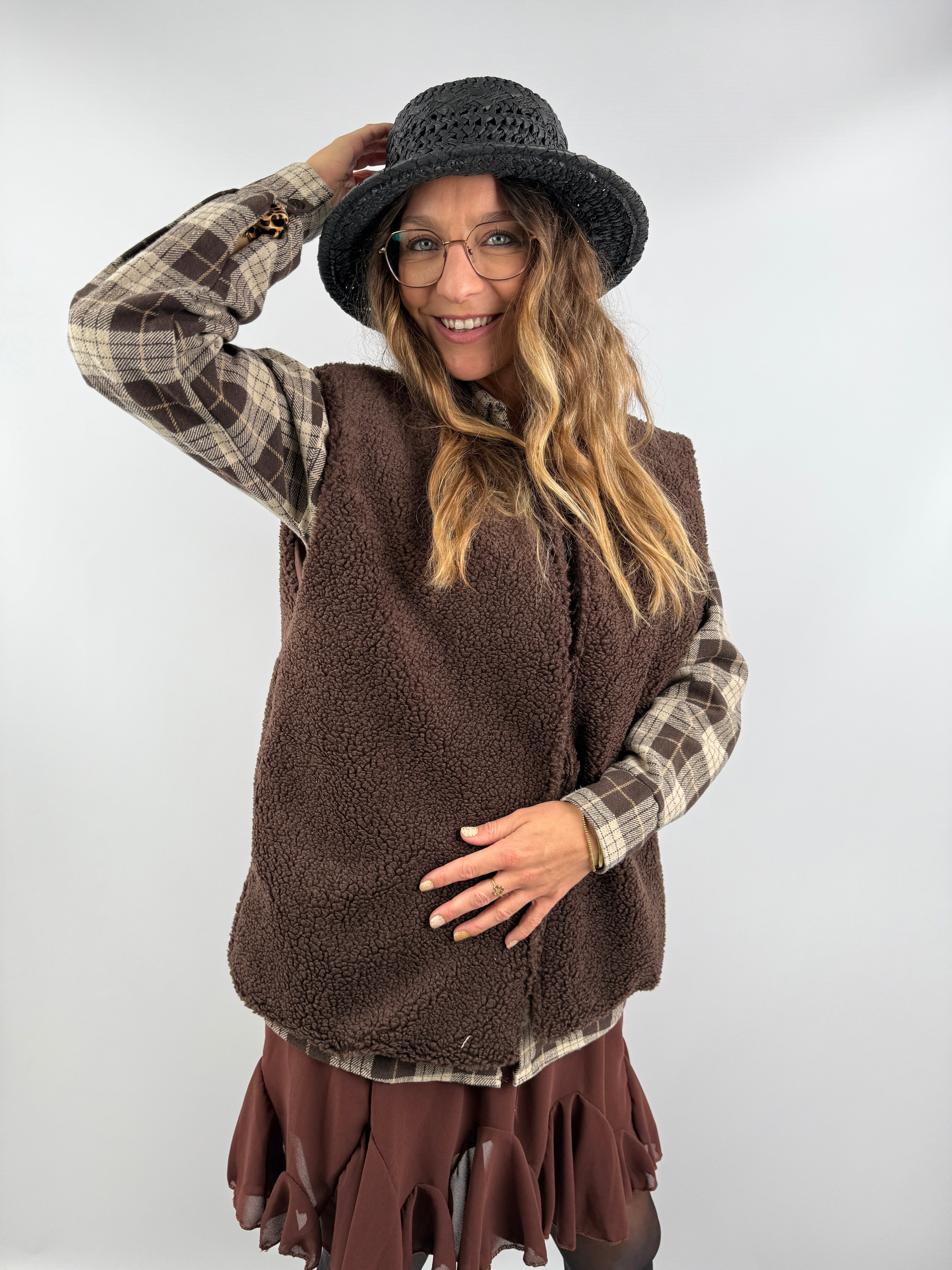 Frau trägt eine braune Teddyweste aus weichem Fleece über einem karierten Overshirt und kombiniert sie mit einem braunen Volant-Rock – gemütlicher Herbstlook mit Stil.
