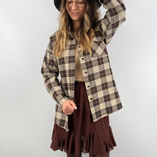 Frau trägt ein braun-beiges kariertes Overshirt aus weichem Flanell, kombiniert mit einem braunen Volant-Rock und schwarzem Hut – herbstlich-femininer Look.