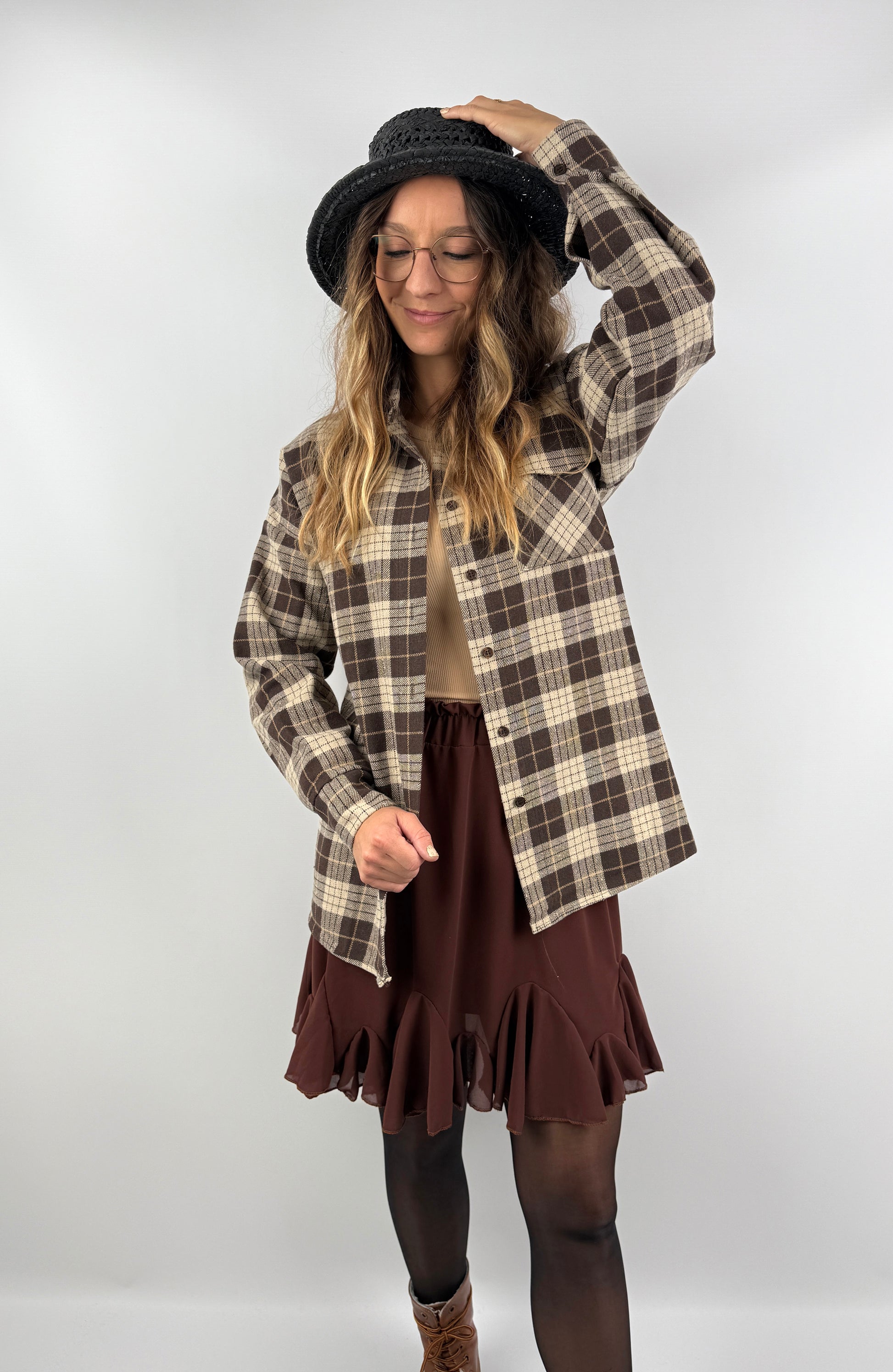 Frau trägt ein braun-beiges kariertes Overshirt aus weichem Flanell, kombiniert mit einem braunen Volant-Rock und schwarzem Hut – herbstlich-femininer Look.