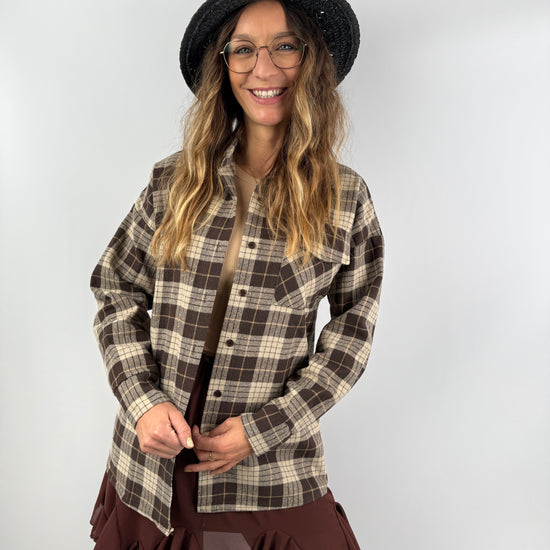 Frau mit kariertem Overshirt in Braun und Beige, kombiniert mit Rüschenrock und schwarzem Strohhut – stilvoller Mix aus Casual und feminin.