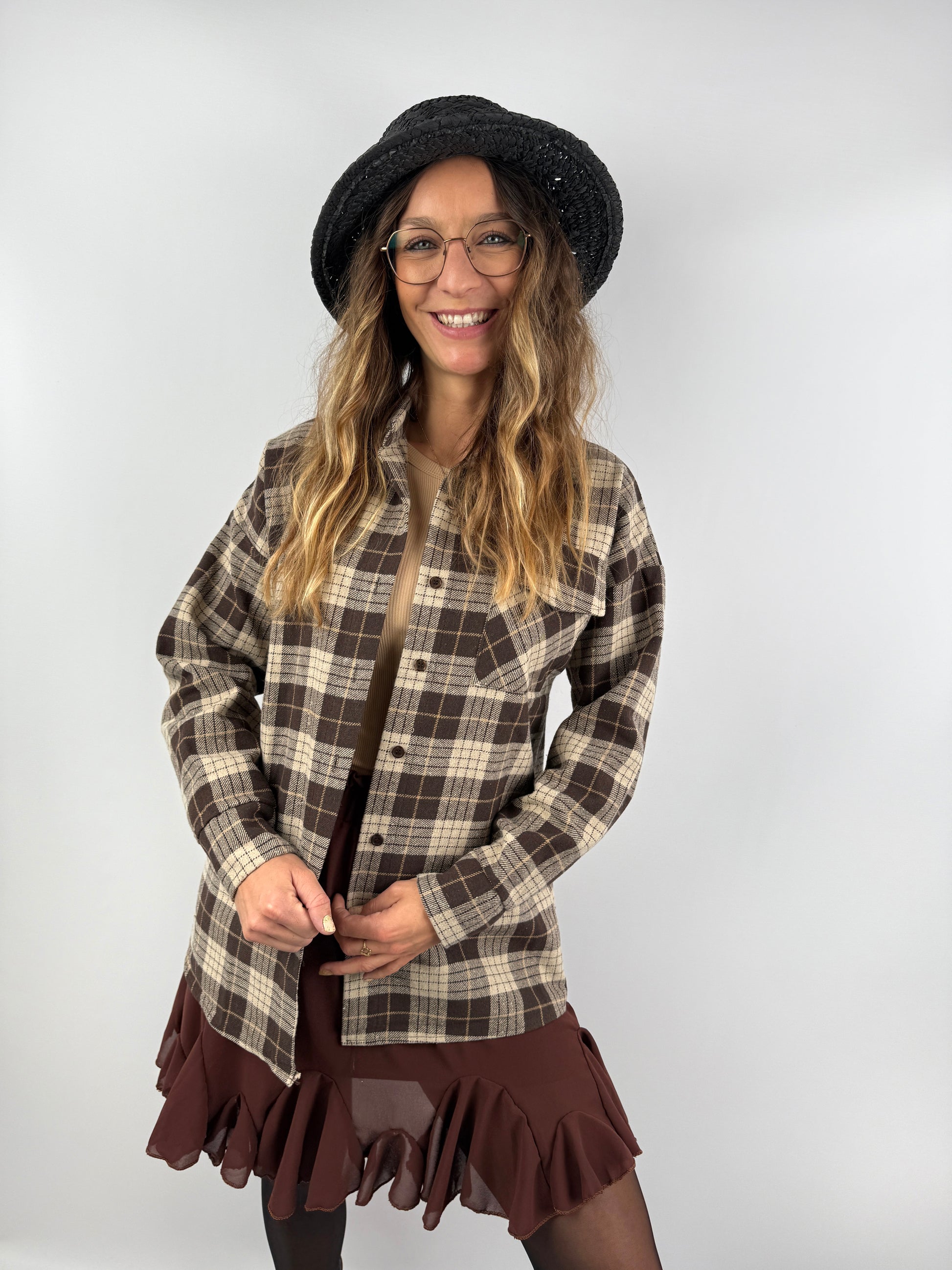 Frau mit kariertem Overshirt in Braun und Beige, kombiniert mit Rüschenrock und schwarzem Strohhut – stilvoller Mix aus Casual und feminin.