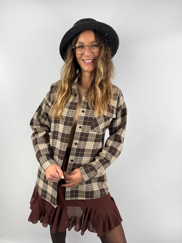 Frau mit kariertem Overshirt in Braun und Beige, kombiniert mit Rüschenrock und schwarzem Strohhut – stilvoller Mix aus Casual und feminin.