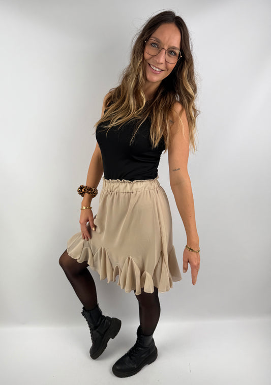 Frau trägt einen beigen Rock mit Volant-Saum und schwarzes Ripp-Top, kombiniert mit schwarzen Boots – moderner, femininer Look mit kontrastreichem Stil.