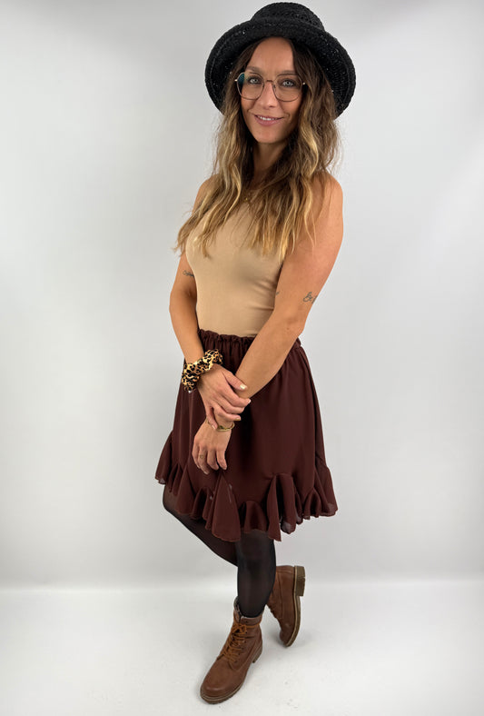 Frau trägt einen braunen Rock mit Volant-Saum, kombiniert mit einem beigefarbenen Ripp-Top, schwarzem Hut und braunen Boots – femininer, herbstlicher Look.