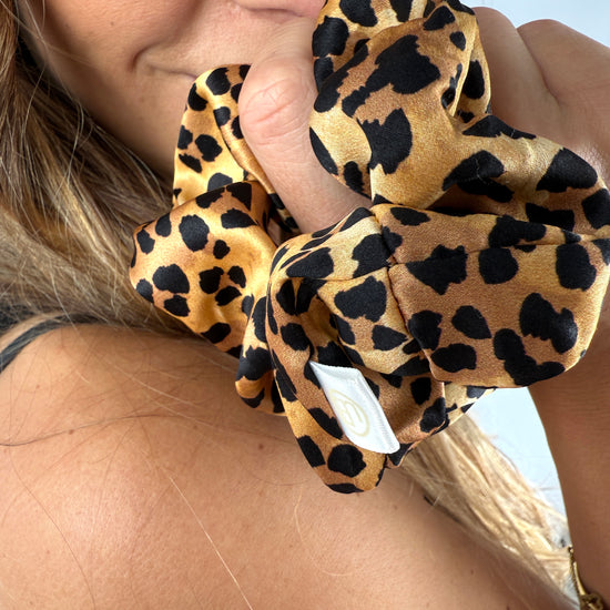 Nahaufnahme eines Seiden-Scrunchies im Leo-Print am Handgelenk – goldene Accessoires ergänzen den Look elegant.
