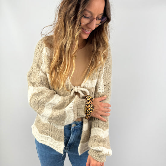 Frau trägt eine flauschige, beige-cremefarbene Strickjacke mit breiten Streifen und Schleifendetail – kombiniert mit Jeans und beigem Top für einen sanften, natürlichen Look.