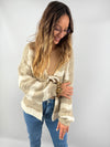 Frau trägt eine flauschige, beige-cremefarbene Strickjacke mit breiten Streifen und Schleifendetail – kombiniert mit Jeans und beigem Top für einen sanften, natürlichen Look.