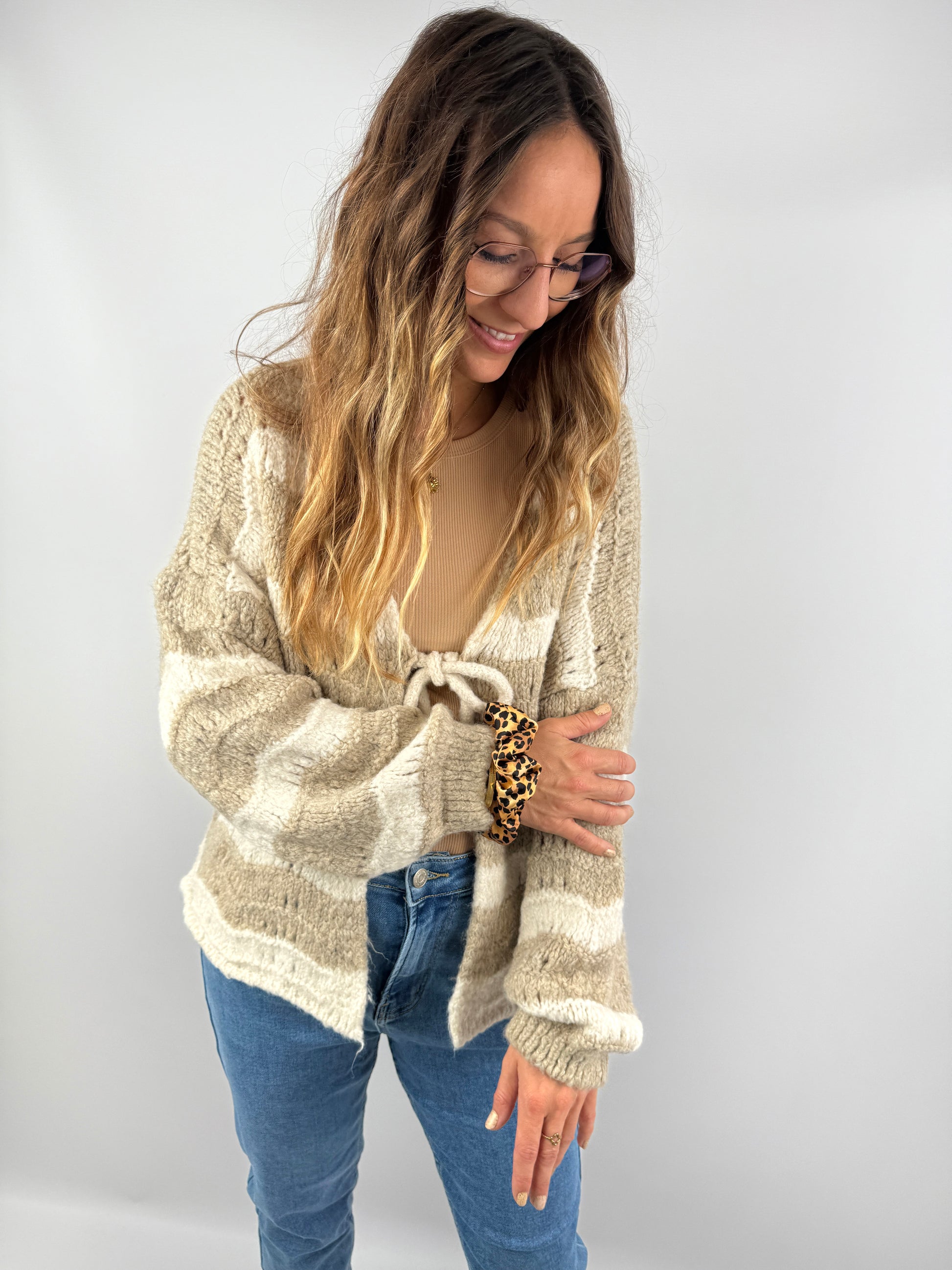 Frau trägt eine flauschige, beige-cremefarbene Strickjacke mit breiten Streifen und Schleifendetail – kombiniert mit Jeans und beigem Top für einen sanften, natürlichen Look.