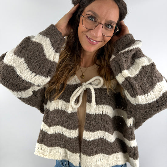 Frau trägt eine braun-cremefarbene Strickjacke mit breiten Streifen und Schleifendetail am Ausschnitt – kuscheliger, femininer Look.