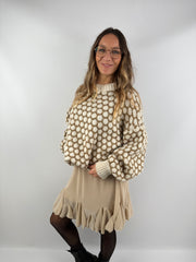 Frau trägt einen oversized beige-cremefarbenen Strickpullover mit 3D-Pünktchenmuster und kombiniert ihn mit einem fließenden beigen Volant-Rock, schwarzer Strumpfhose und Boots.