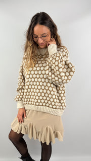Frau trägt einen oversized beige-cremefarbenen Strickpullover mit 3D-Pünktchenmuster und kombiniert ihn mit einem fließenden beigen Volant-Rock, schwarzer Strumpfhose und Boots.
