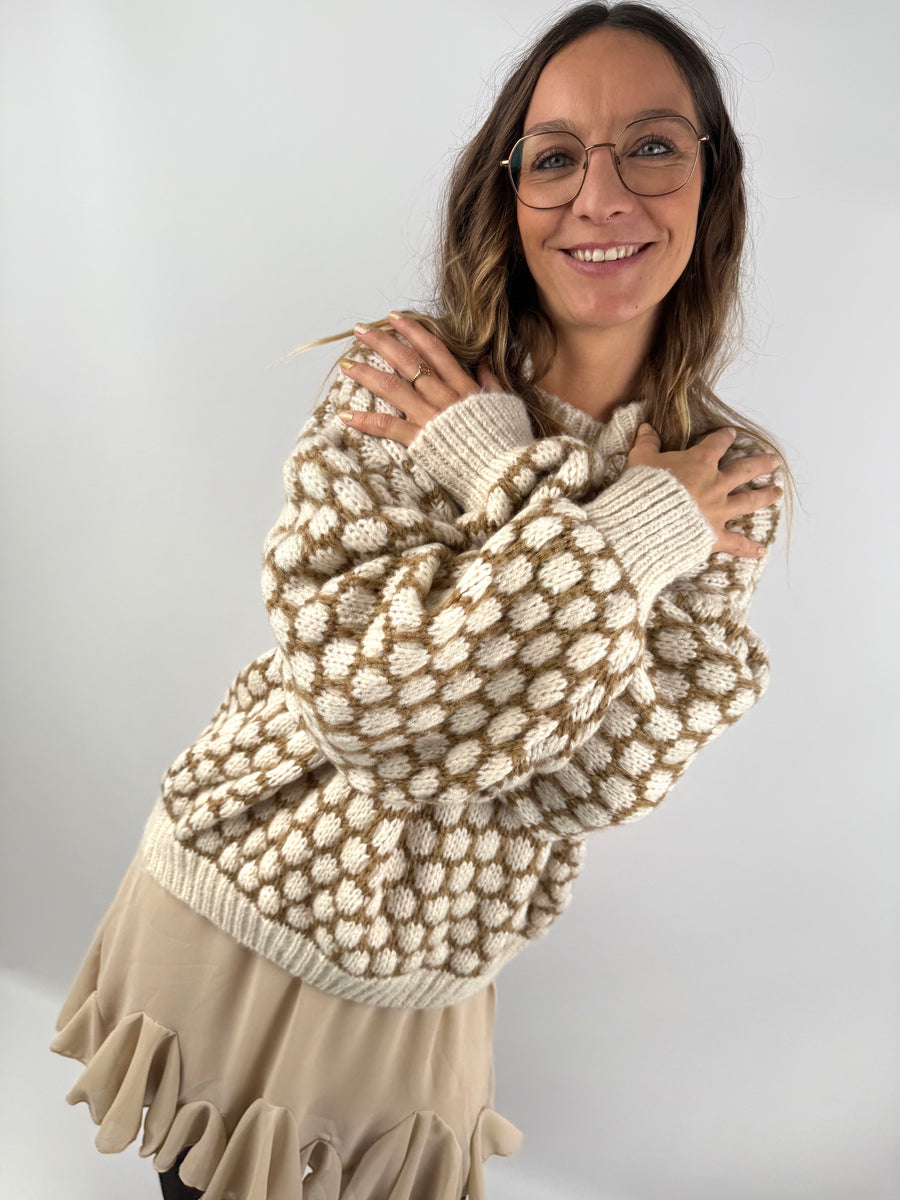 Frau trägt einen oversized beige-cremefarbenen Strickpullover mit 3D-Pünktchenmuster und kombiniert ihn mit einem fließenden beigen Volant-Rock, schwarzer Strumpfhose und Boots.