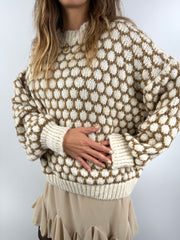 Frau trägt einen oversized beige-cremefarbenen Strickpullover mit 3D-Pünktchenmuster und kombiniert ihn mit einem fließenden beigen Volant-Rock, schwarzer Strumpfhose und Boots.