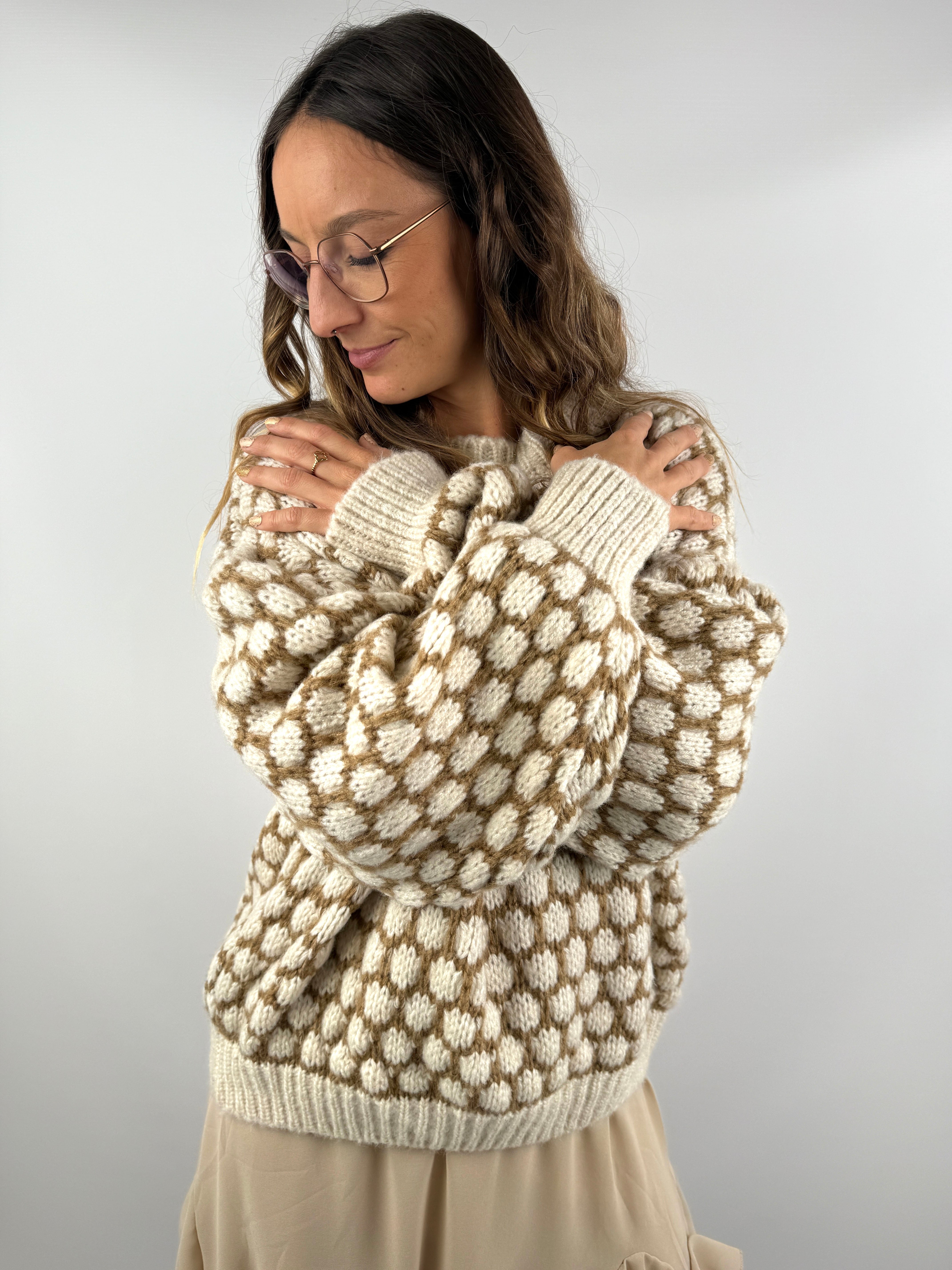 Frau trägt einen oversized beige-cremefarbenen Strickpullover mit 3D-Pünktchenmuster und kombiniert ihn mit einem fließenden beigen Volant-Rock, schwarzer Strumpfhose und Boots.