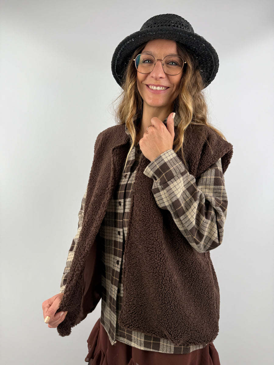 Frau trägt eine braune Weste aus Teddyfell über einem karierten Hemd – moderner, kuscheliger Look mit femininen Details.
